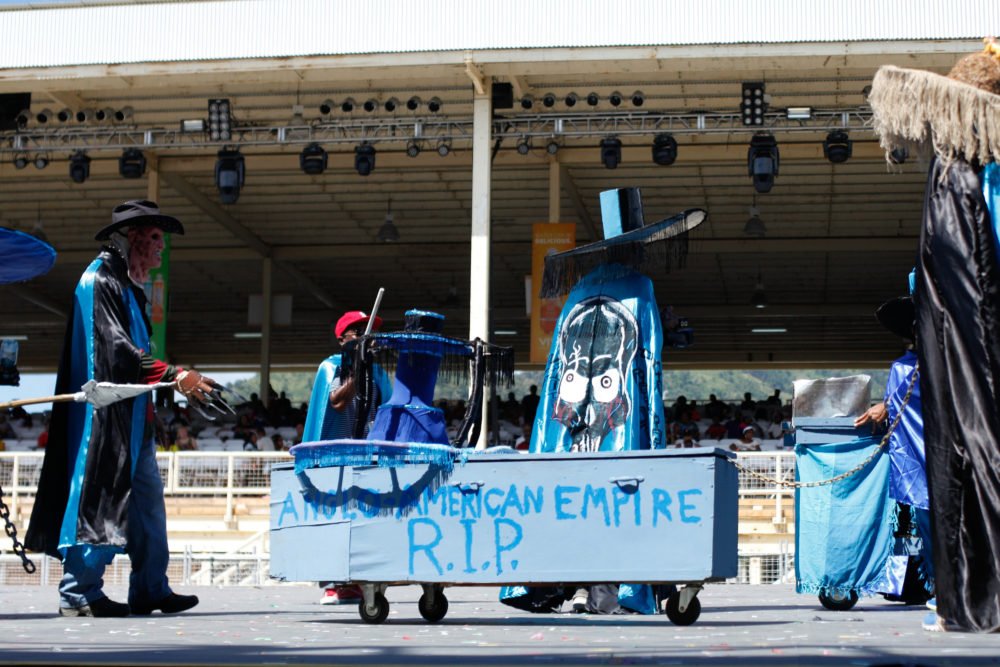 Part of a political commentary mas band, pulling a coffin that says "Anglo-American Empire R.I.P. "